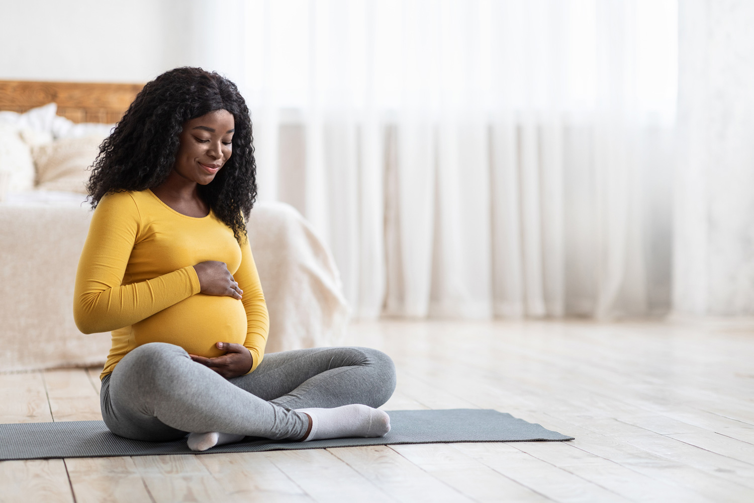 A pregnant woman is sitting on a yoga mat in her bedroom.