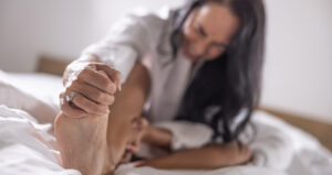 A person lying on a bed is stretching their foot with a focused expression, while wearing a ring on their finger.
