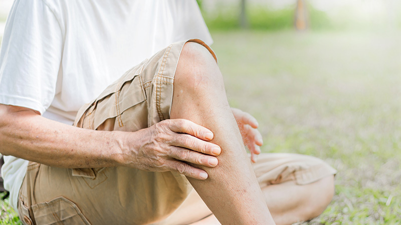 An older adult sits outdoors on grass, holding their lower leg and knee with both hands, possibly indicating chronic venous insufficiency.