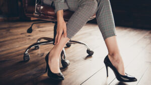 A person sitting on an office chair touches their ankle while wearing plaid pants and black high-heeled shoes on a wooden floor.
