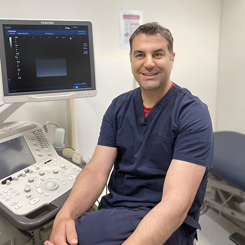 A man in medical scrubs sits next to an ultrasound machine in a clinical examination room, smiling at the camera.